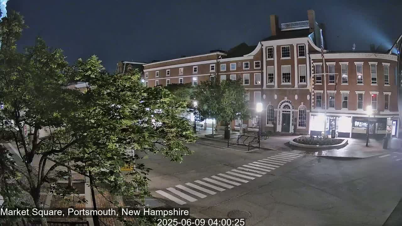 A nighttime view of a mostly empty town square with brick buildings and trees under a clear, dark sky.