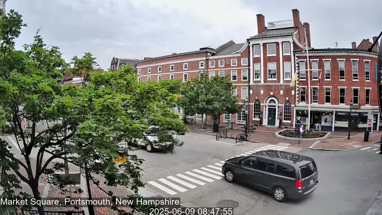 A brick-building-lined street intersection with pedestrian crosswalks, several vehicles, and lush green trees under an overcast sky.