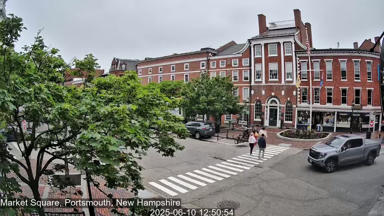 A pedestrian crosswalk in a town square with several people walking across it, under a cloudy sky, features brick buildings, lush green trees, and parked and moving cars.