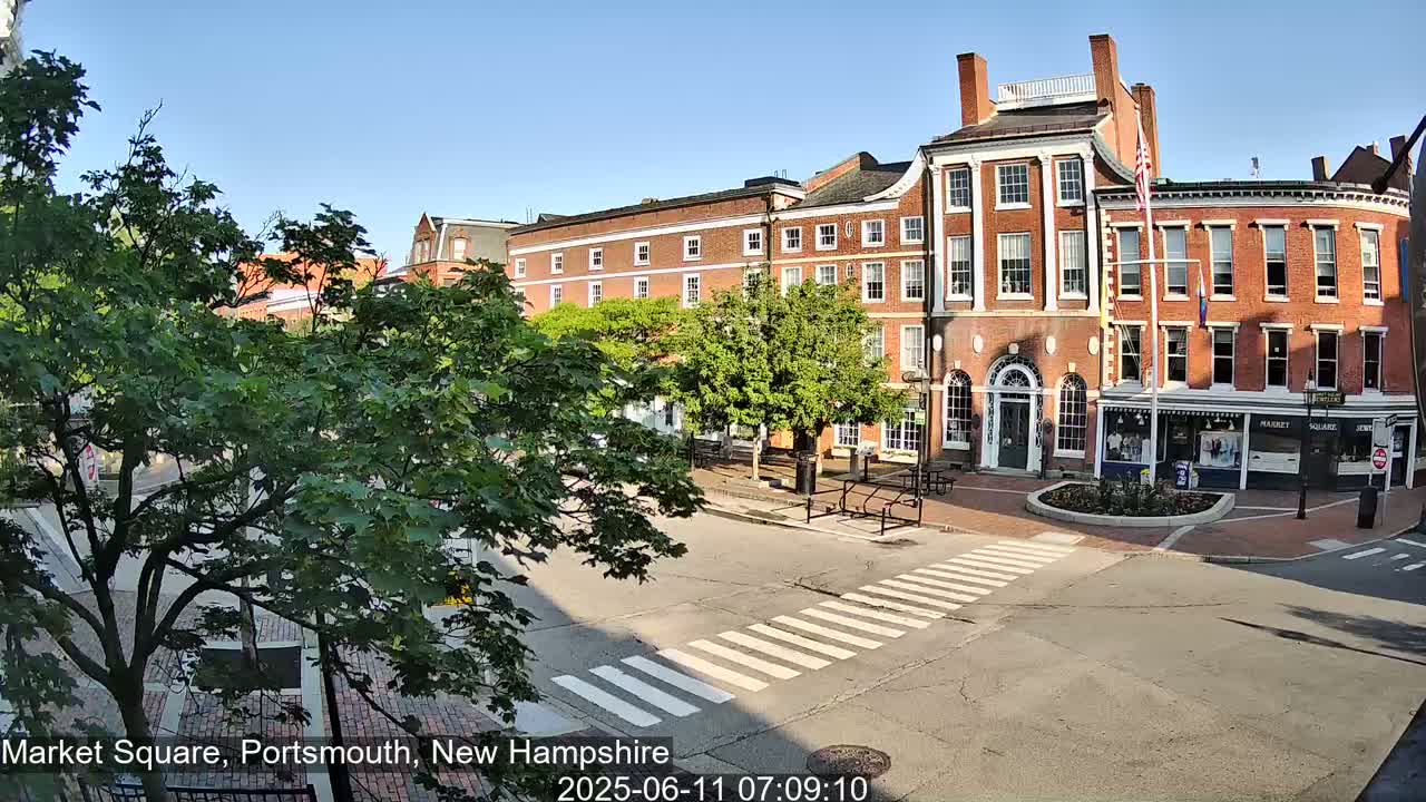 A sunny day reveals a brick-building-lined town square with a pedestrian crosswalk, and leafy green trees.