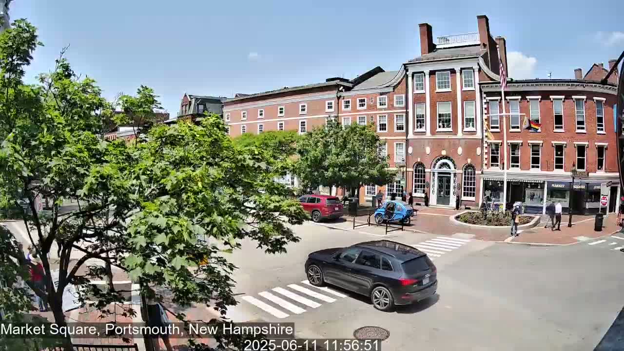 A sunny day in a city square shows several brick buildings, green trees, pedestrians, and cars at a crosswalk.