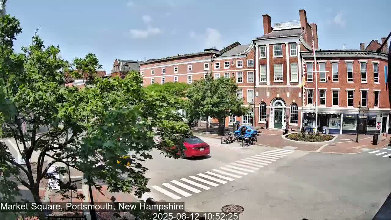 A sunny day in a brick-lined town square shows several buildings, a pedestrian crossing, some parked bicycles, and a red car driving through the intersection.