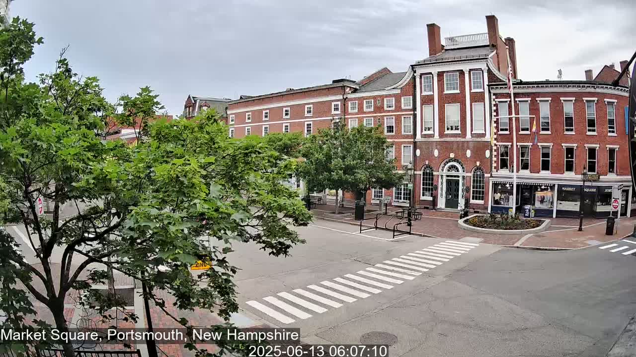 A brick building in a town square on an overcast day, with leafy green trees and a pedestrian crossing in the foreground.