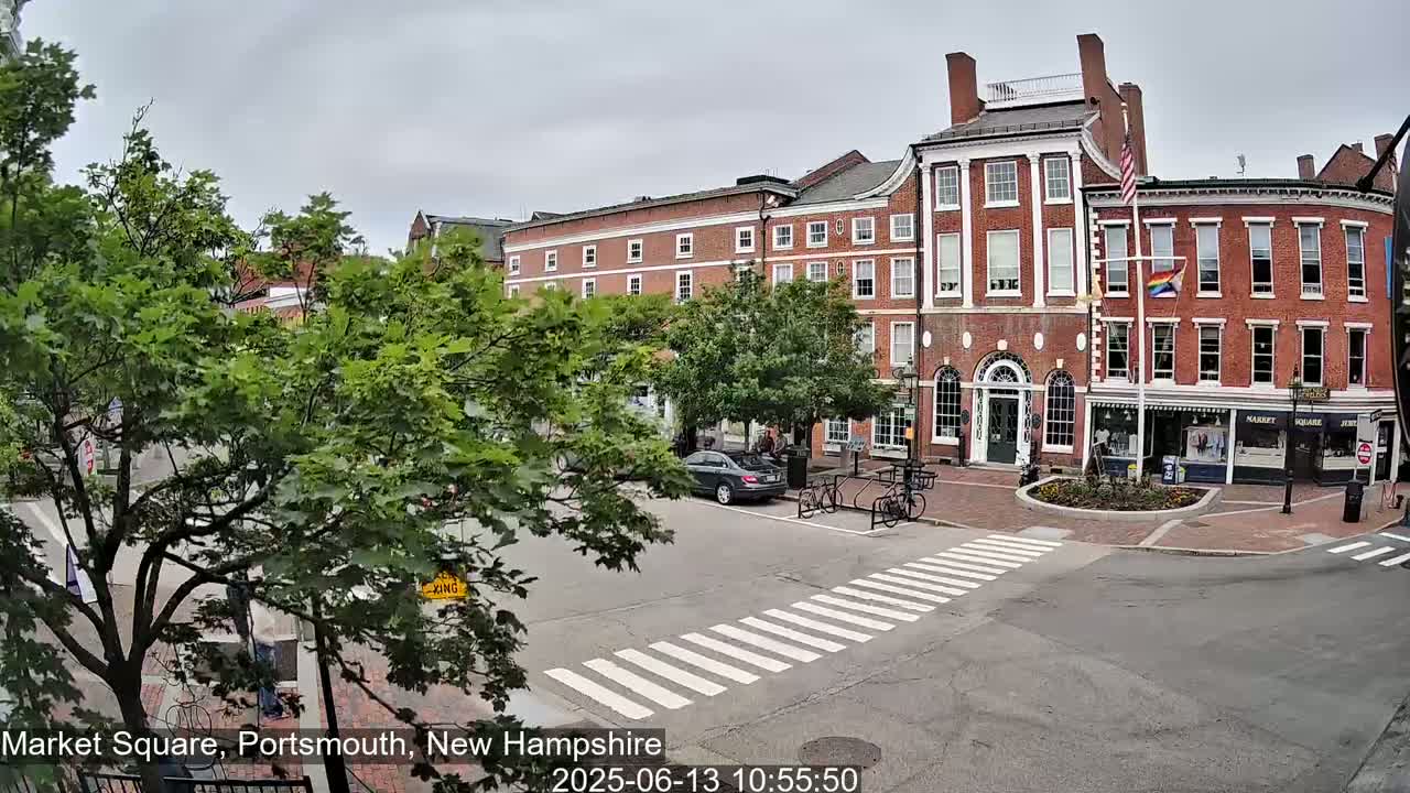A brick building with shops on the ground floor sits in a town square, with overcast skies and cars parked nearby.