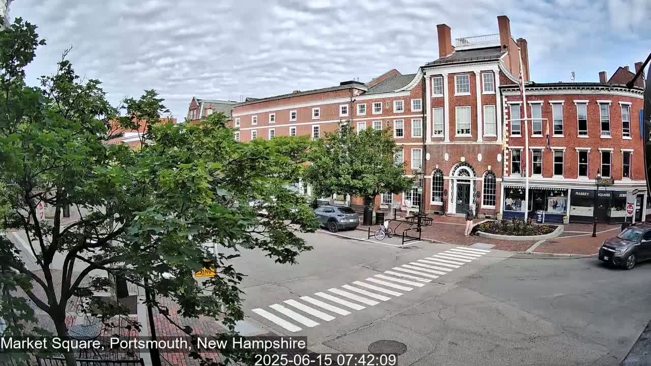 A partly cloudy day reveals a brick building complex in a town square, with pedestrians crossing a crosswalk and cars parked and driving nearby.