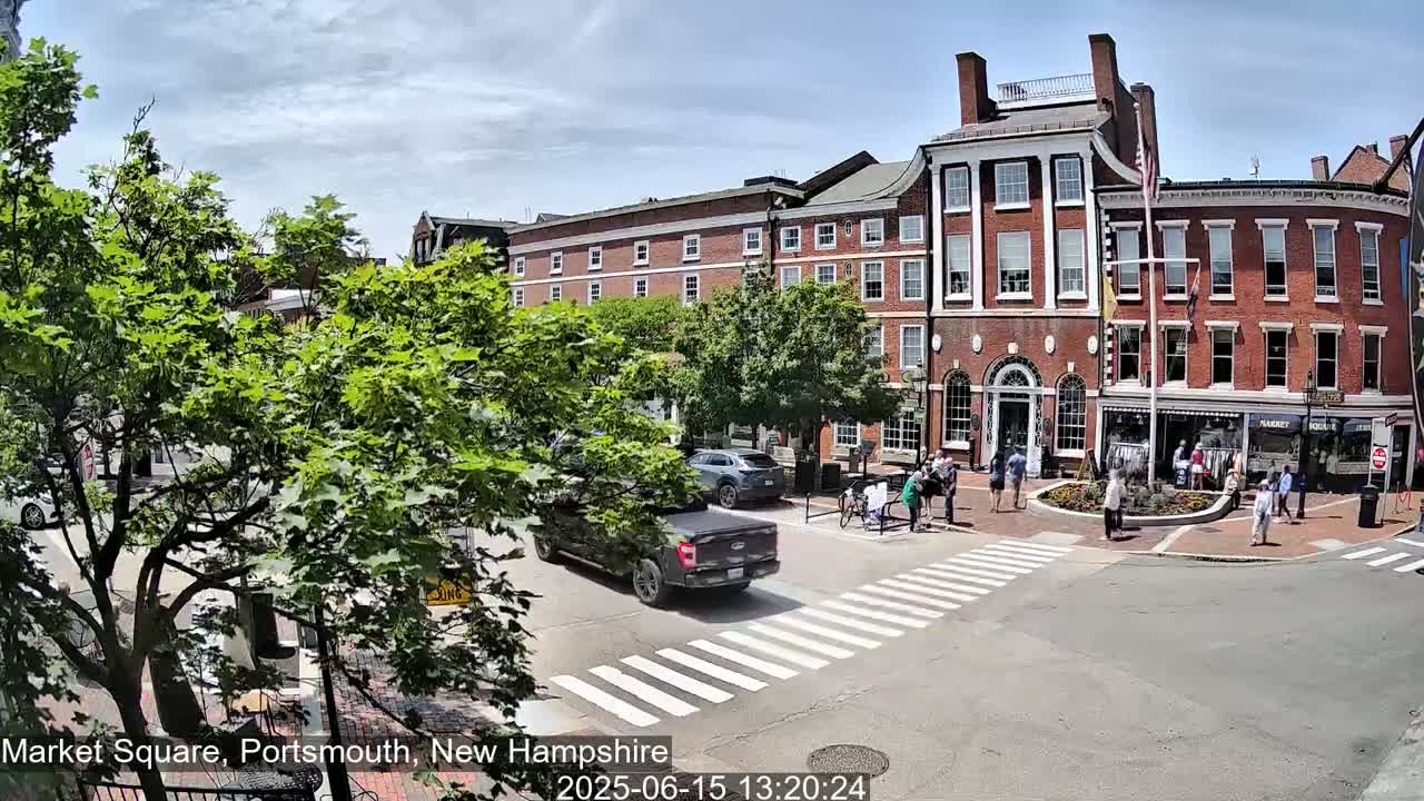A sunny day in a city square shows pedestrians crossing a street in front of several brick buildings, with cars parked and driving nearby, and lush green trees lining the streets.