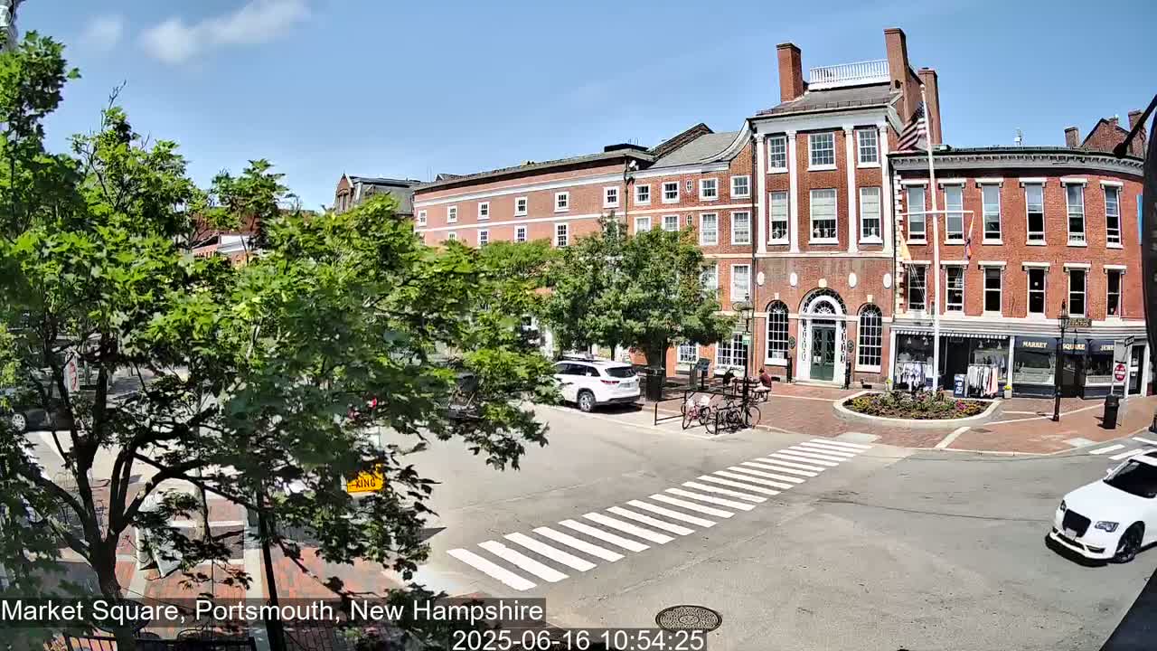 A sunny day in a brick-building town square shows a pedestrian crossing, parked cars, and several people sitting at outdoor tables.