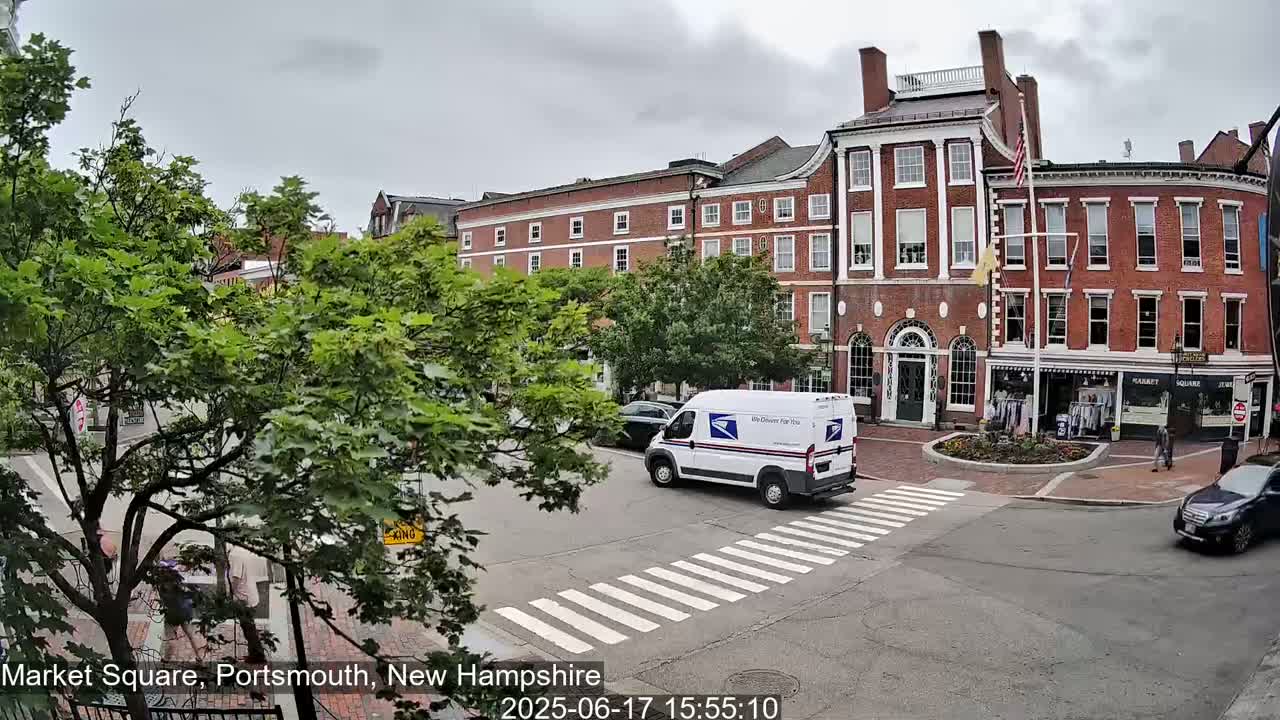 A postal truck drives through a tree-lined intersection in front of brick buildings under an overcast sky.