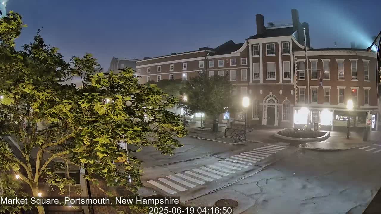 A nighttime, slightly foggy view of a town square with brick buildings, streetlights, and lush green trees.