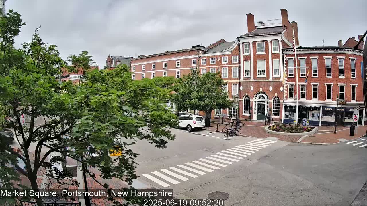 A partly cloudy day shows a brick-fronted town square with parked cars and a crosswalk.