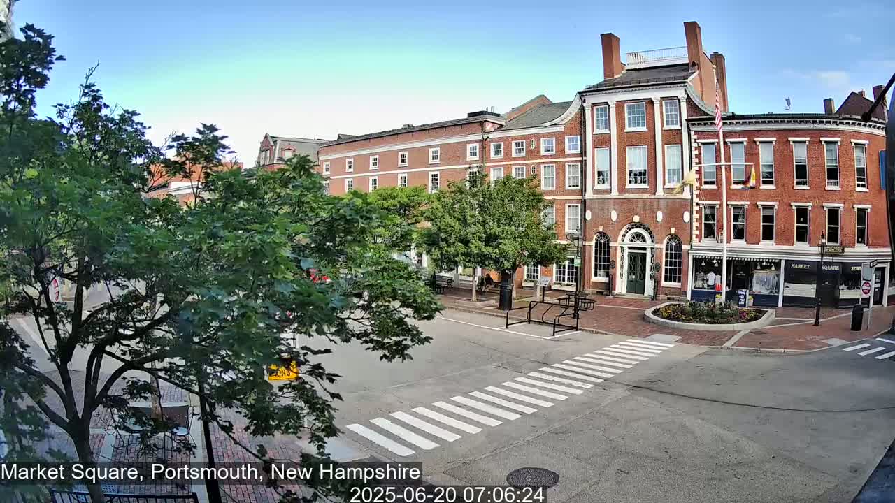 A sunny view of a brick building and town square with a crosswalk and lush green trees.