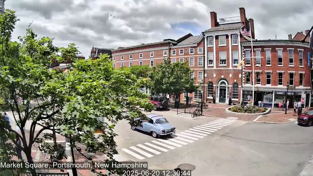 A partly cloudy day shows a quaint town square with several brick buildings, lush green trees, a pedestrian crossing, and a few cars, including a light blue vintage car.