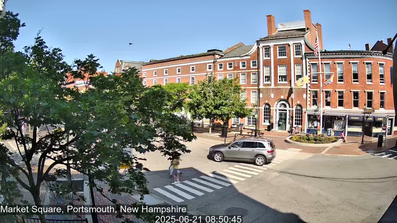 A sunny day reveals a city square with brick buildings, green trees, and a silver SUV crossing a pedestrian crossing.