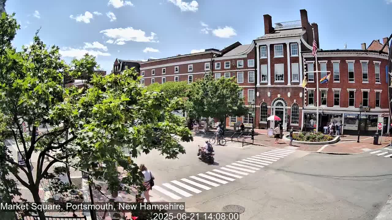 A sunny day in a town square shows a brick building, pedestrians crossing a street, and several people on bicycles and a scooter.