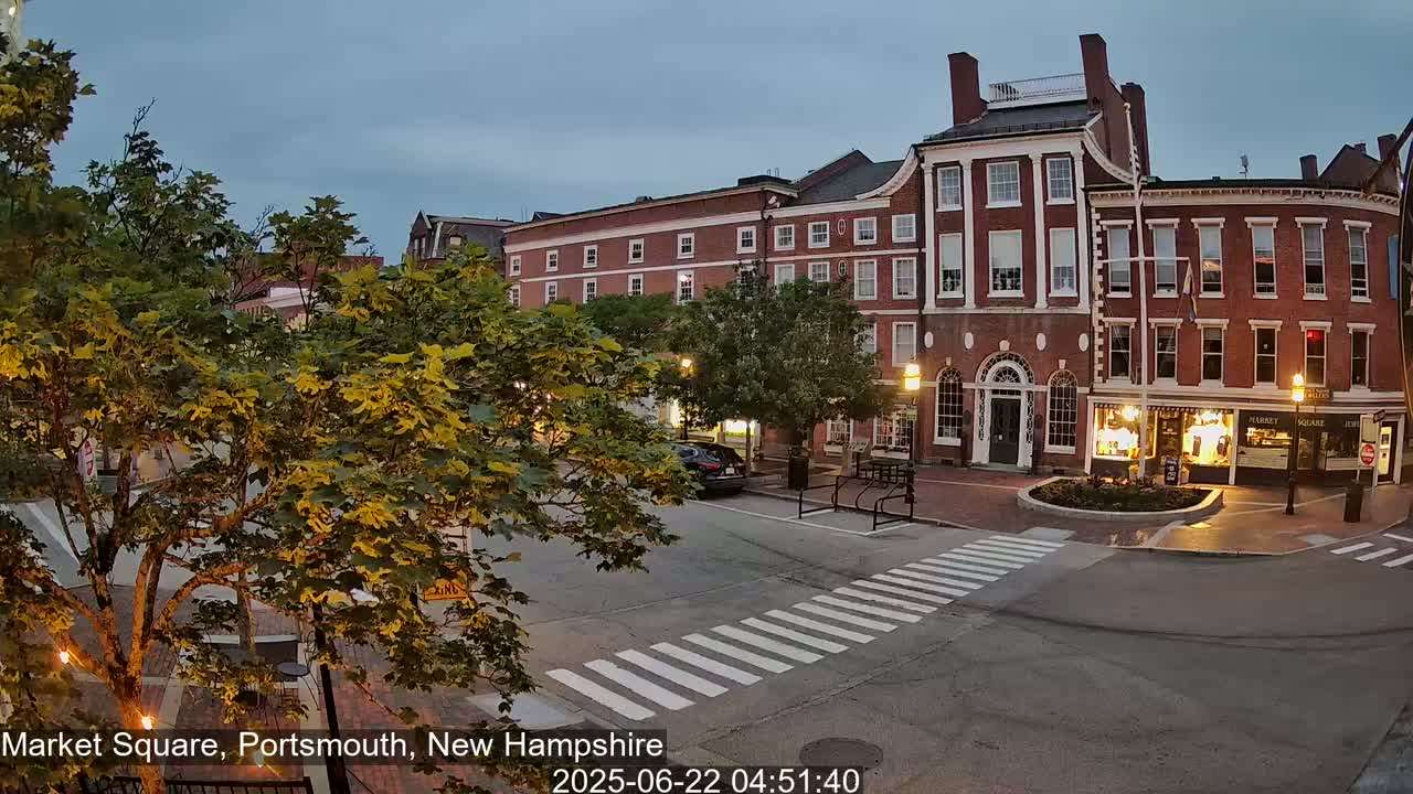 A mostly empty town square at twilight, with brick buildings and lush green trees lining the streets.