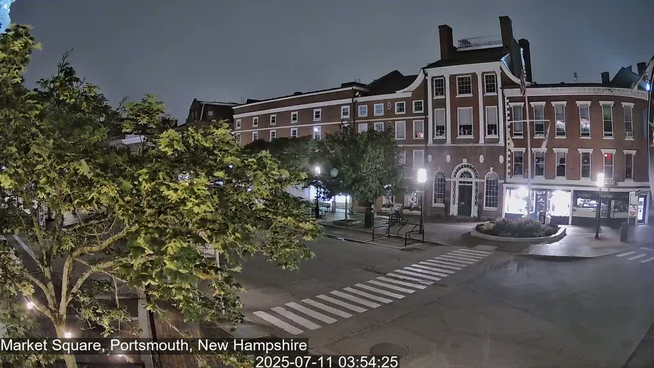 A nighttime view of a mostly empty town square with brick buildings, green trees, and a crosswalk.