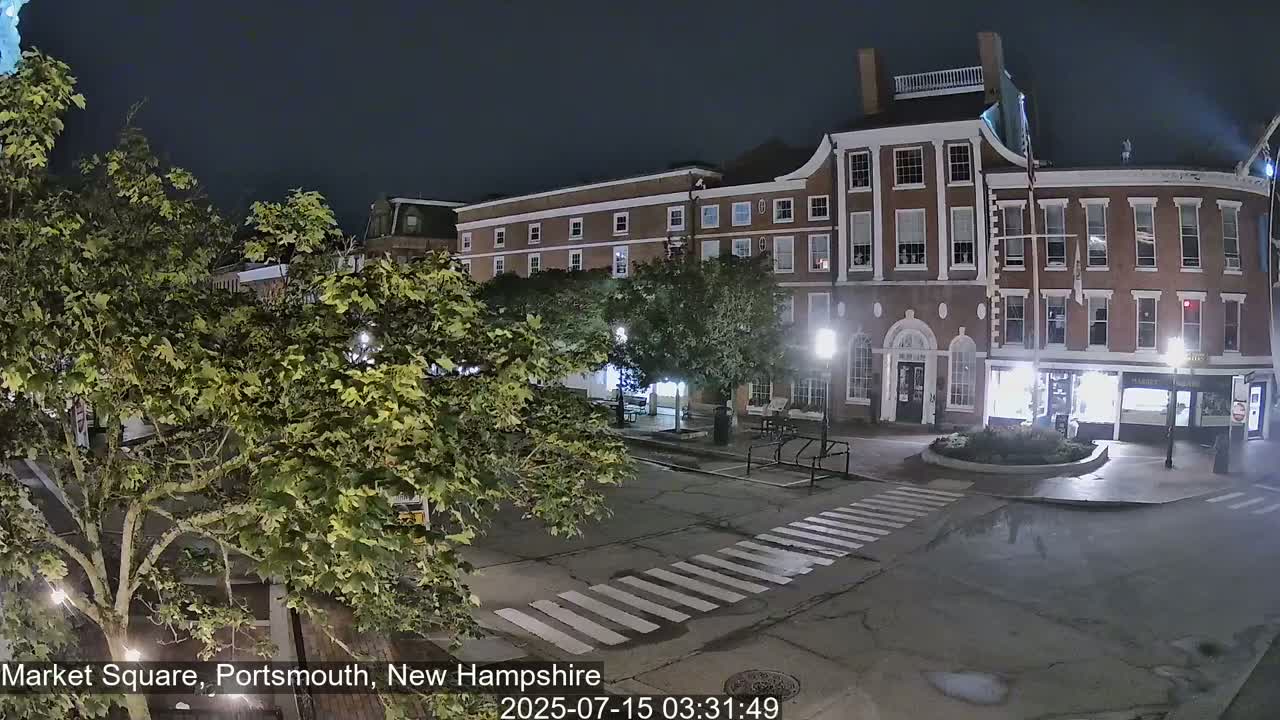 A nighttime view of a brick building in a town square, with leafy trees and a crosswalk visible in the foreground under dark skies.