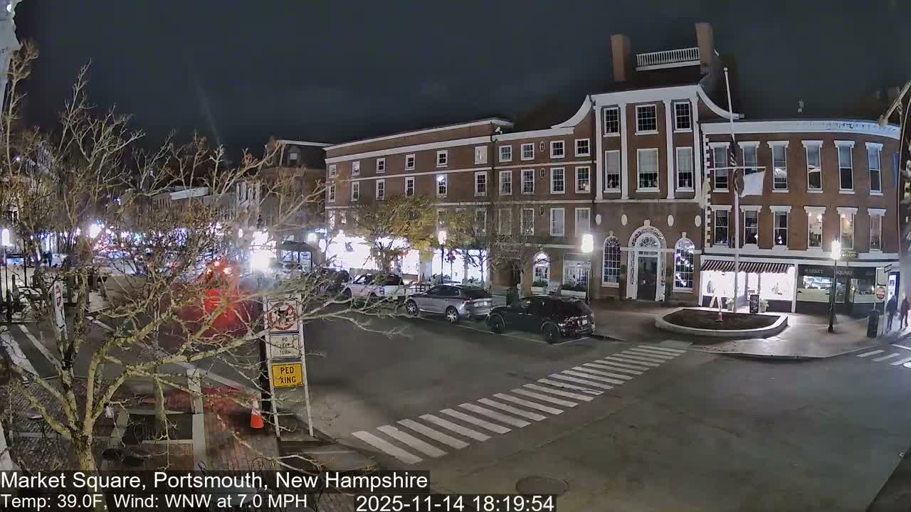 The image captures a chilly, clear night in a bustling city square, featuring historic brick buildings illuminated by streetlights, parked cars lining the street, bare trees, and a crosswalk with a few pedestrians.