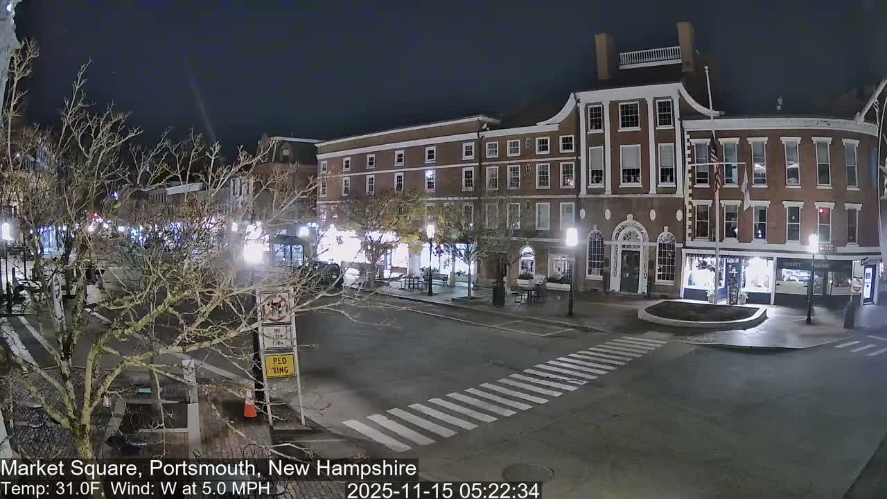 A quiet, well-lit urban square is depicted at night, featuring bare trees, multi-story brick buildings with illuminated windows, and an empty crosswalk under a clear, dark sky.