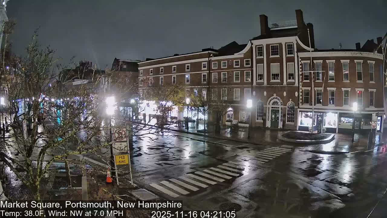 A wet, reflective street with a prominent crosswalk is illuminated by numerous streetlights, surrounded by dark, multi-story brick buildings and bare trees in a town square on an overcast night.