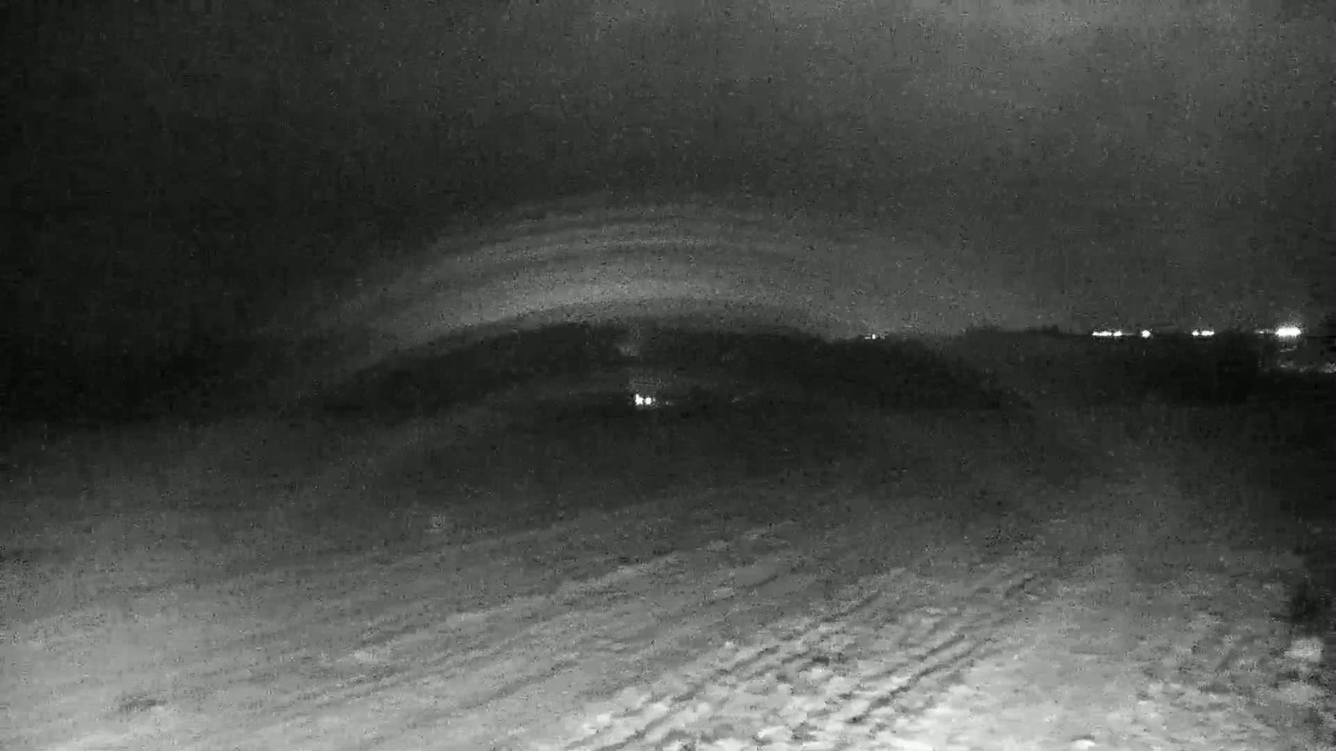 A dark, snowy landscape at night is illuminated by a faint, arched glow in the sky and distant lights.