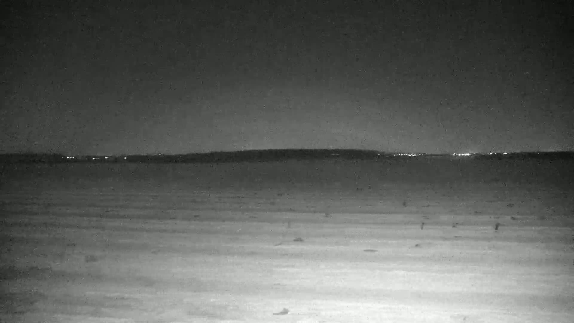A dark, flat expanse of land stretches to a distant horizon where faint lights illuminate a dark landmass under a nighttime sky.