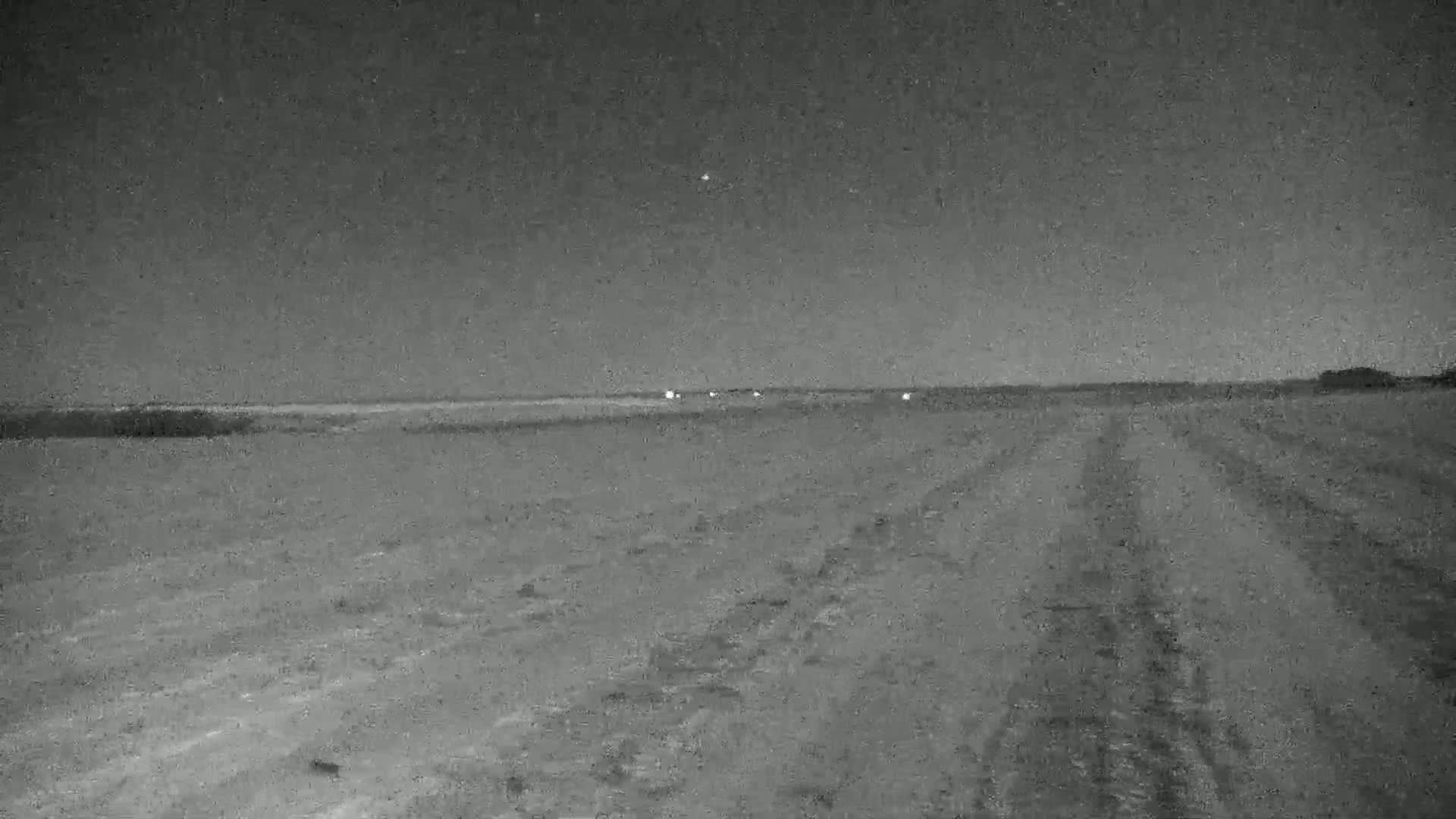A flat, dark landscape at night shows tire tracks leading toward faint lights in the distance under a clear, starry sky.