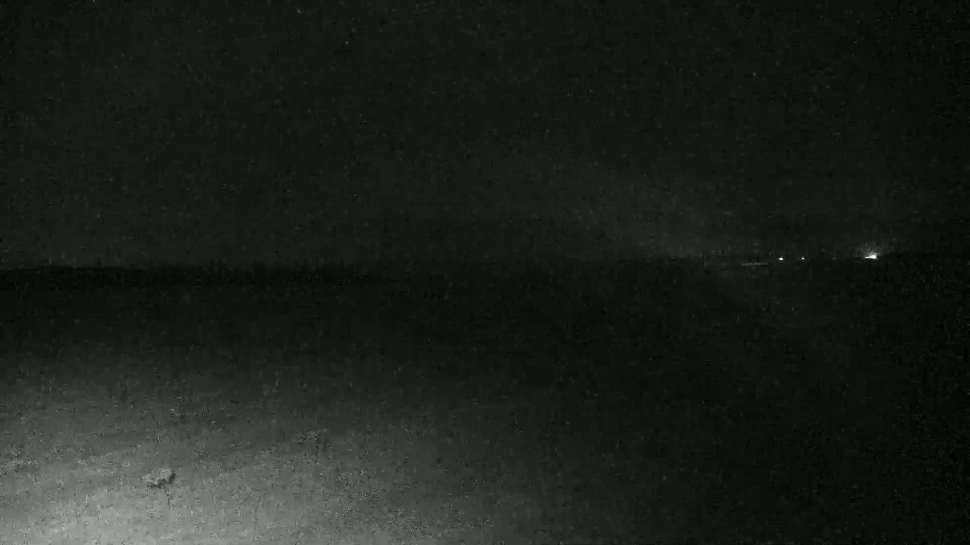 A dark, nighttime scene shows a flat, dark landscape with a few distant lights under a clear, star-filled sky.