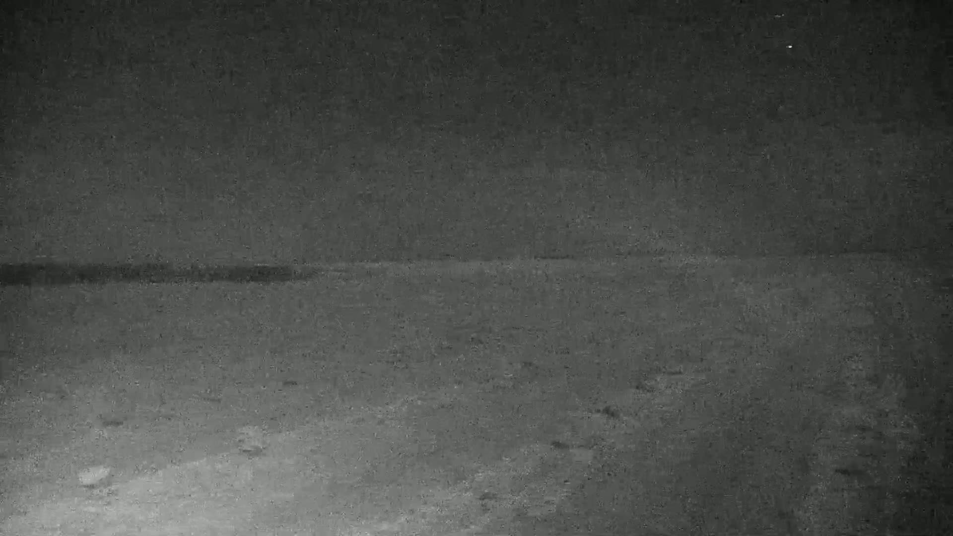 A dark, possibly nighttime, image shows a flat expanse of ground with a slightly darker horizontal line in the middle distance.
