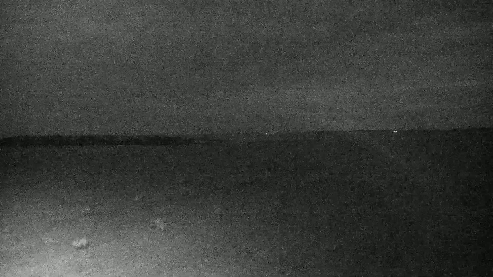 A low-light, nighttime image shows a dark, flat landscape with a few distant lights on the horizon under a mostly clear sky.