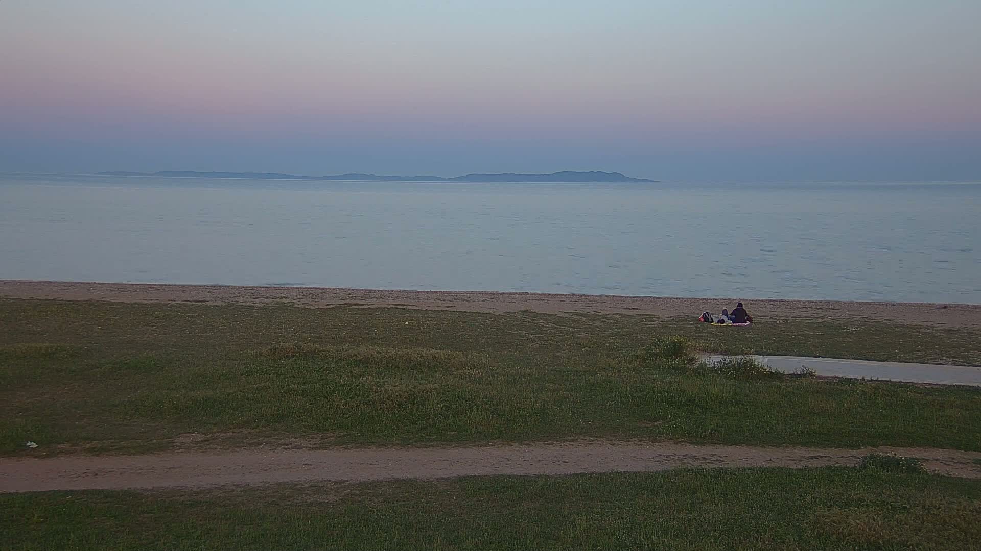 Two people sit on a blanket on a grassy area overlooking a calm sea and a distant landmass under a pale, twilight sky.