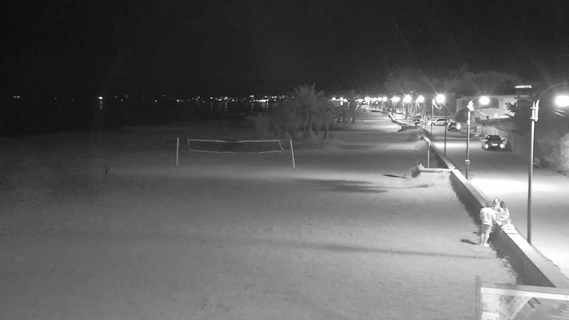 A nighttime view of a beach with a volleyball net, palm trees, and a paved walkway lined with streetlights.
