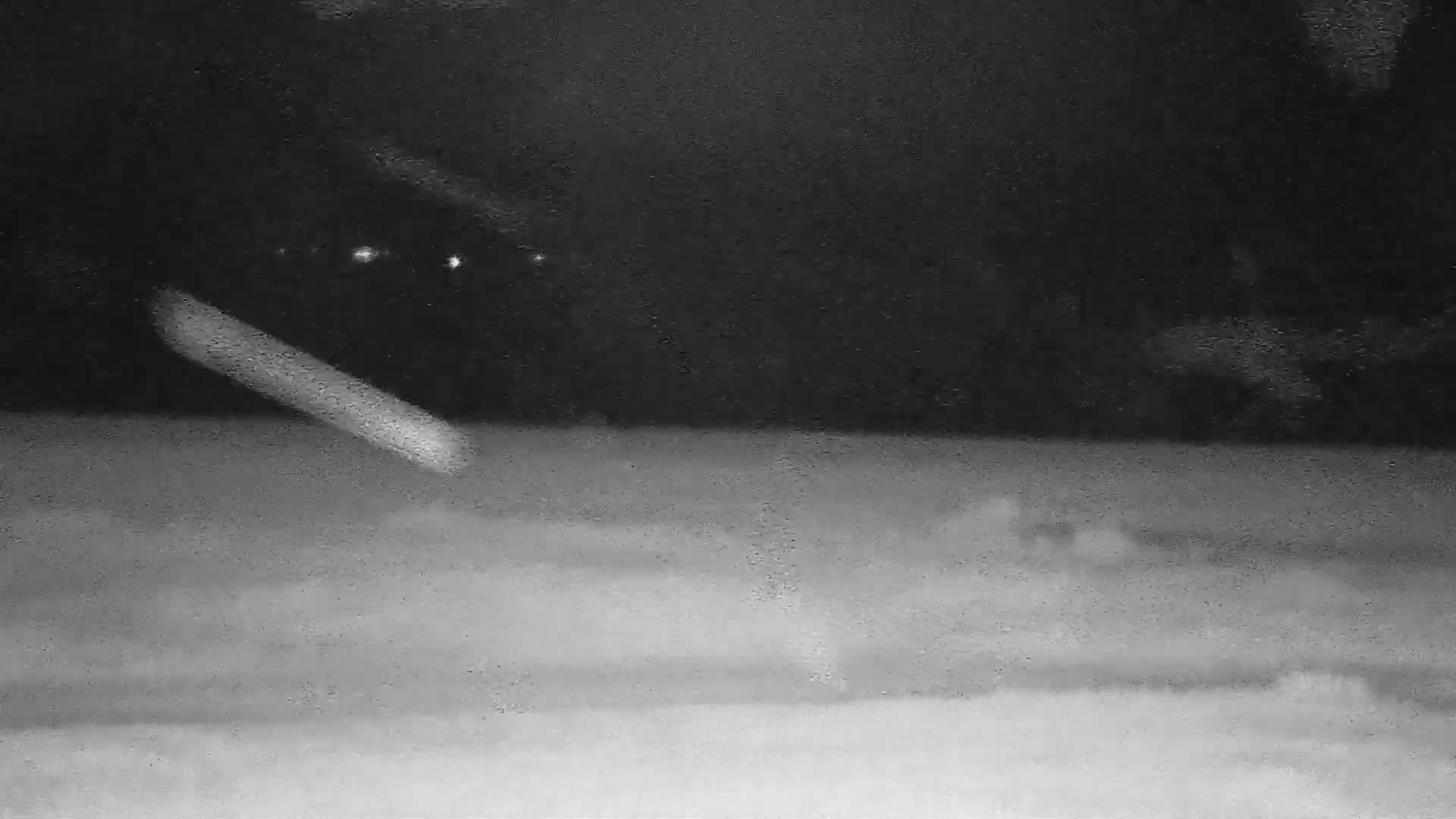 A blurry, grainy nighttime image shows a bright streak of light descending diagonally across a dark sky toward a lighter-colored ground.