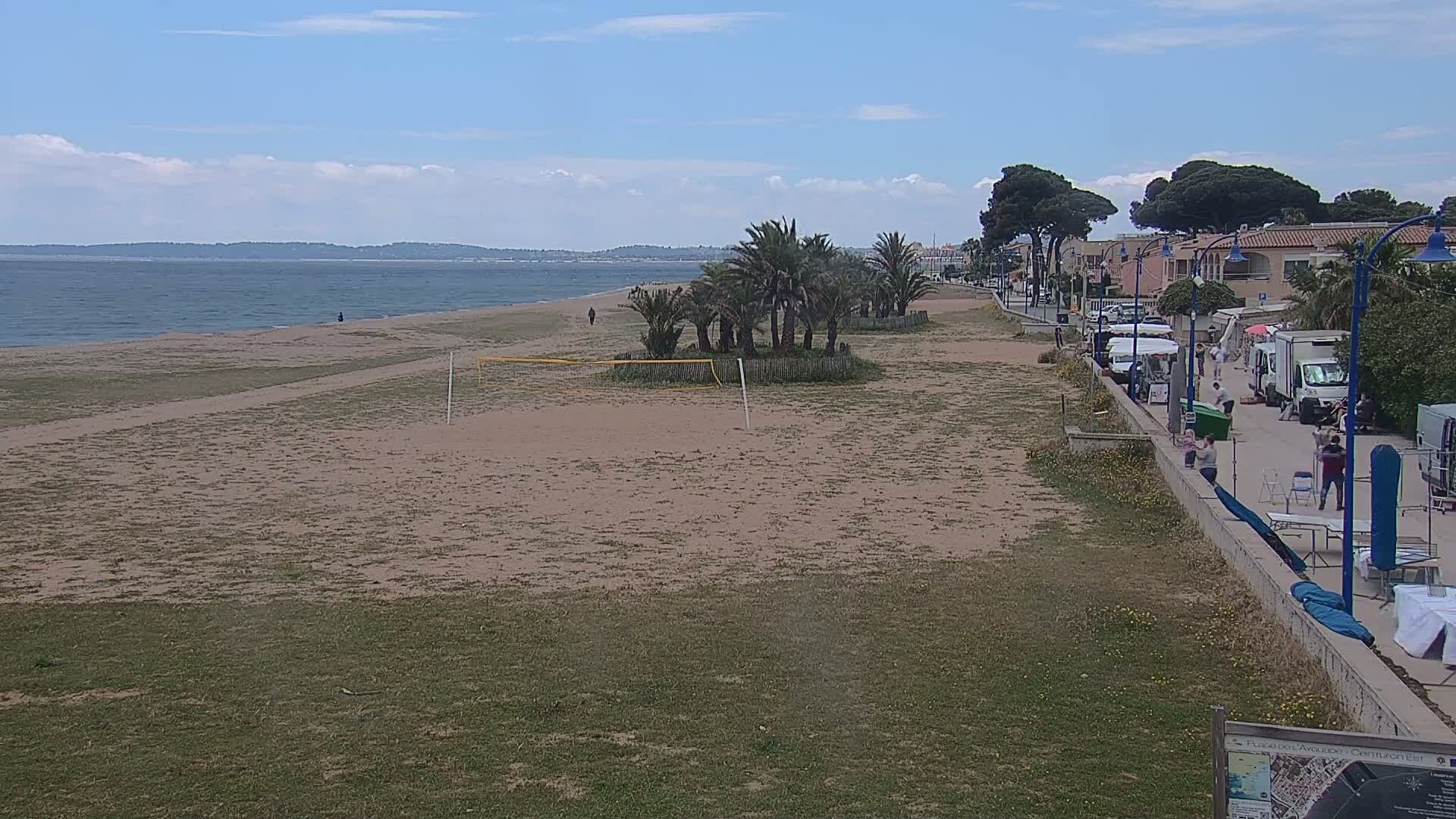 A sandy beach with a volleyball net, palm trees, and buildings along a paved walkway stretches along a calm sea under a partly cloudy sky.