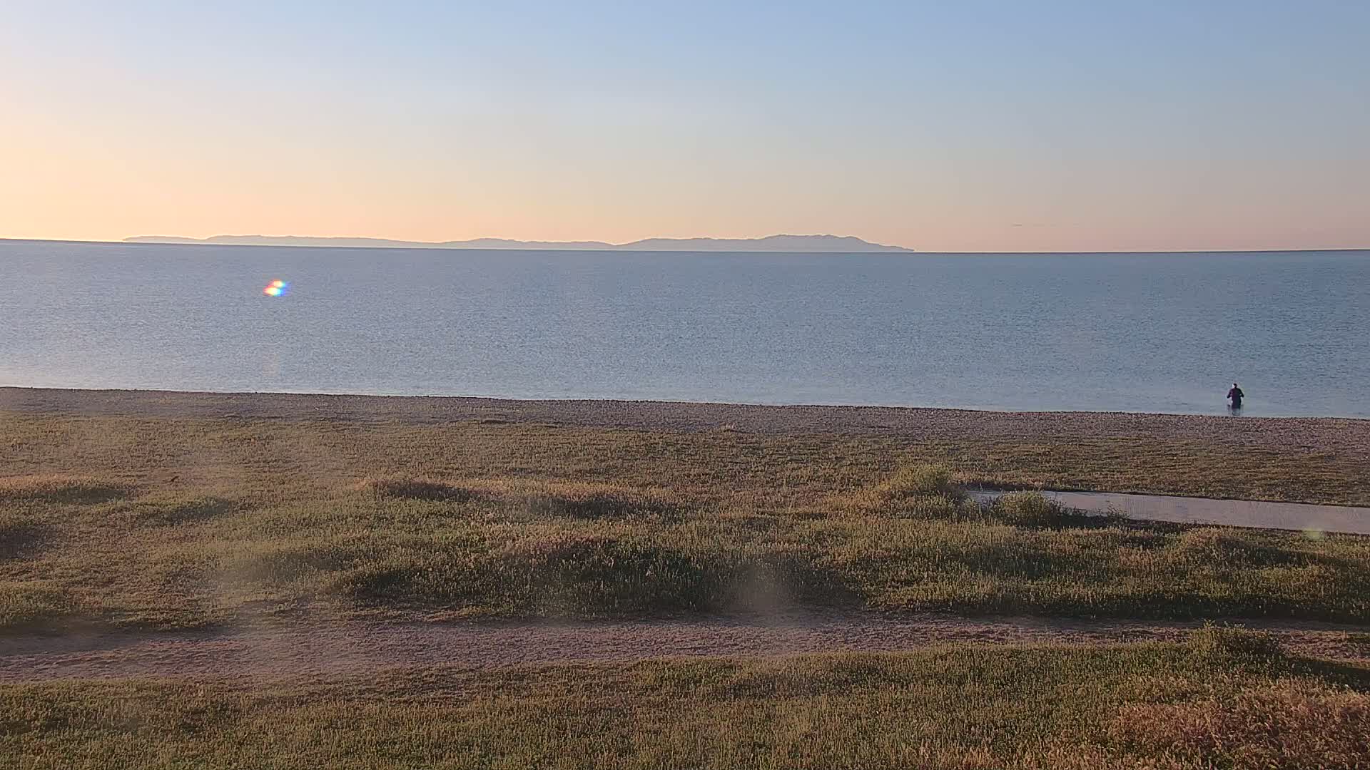 A person stands in shallow water near a beach with grassy dunes, overlooking a calm sea and a distant mountain range under a clear, sunset sky.