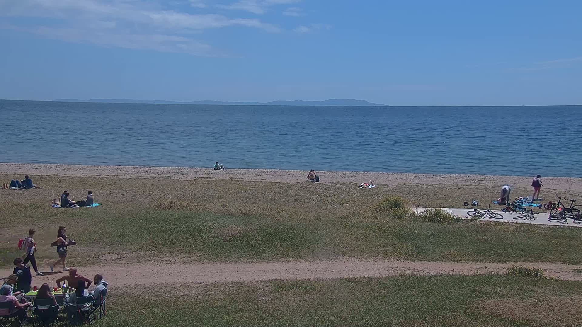 A sandy beach with people sunbathing, picnicking, and riding bicycles, overlooks a calm blue ocean under a mostly sunny sky.