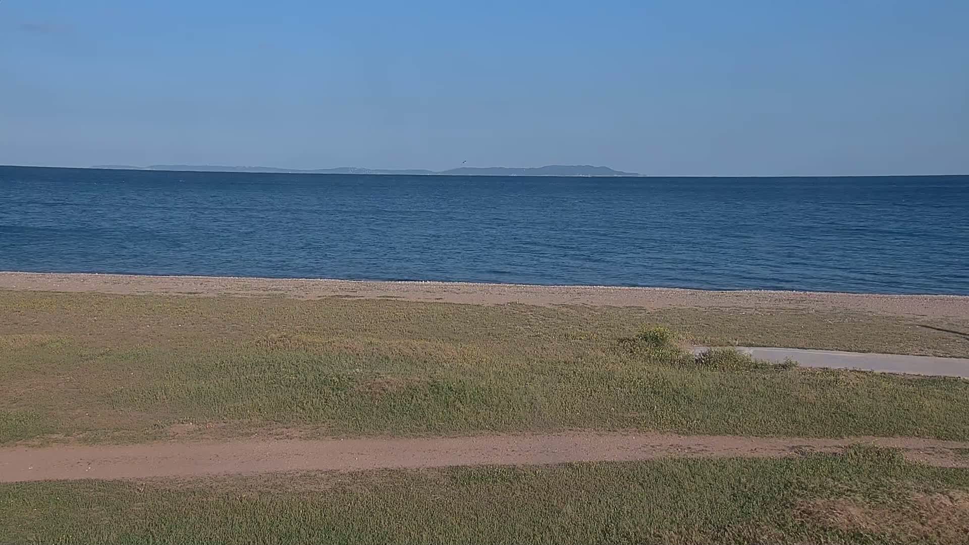 A calm, blue ocean stretches to a distant island under a clear sky, with a grassy area and a sandy beach in the foreground.