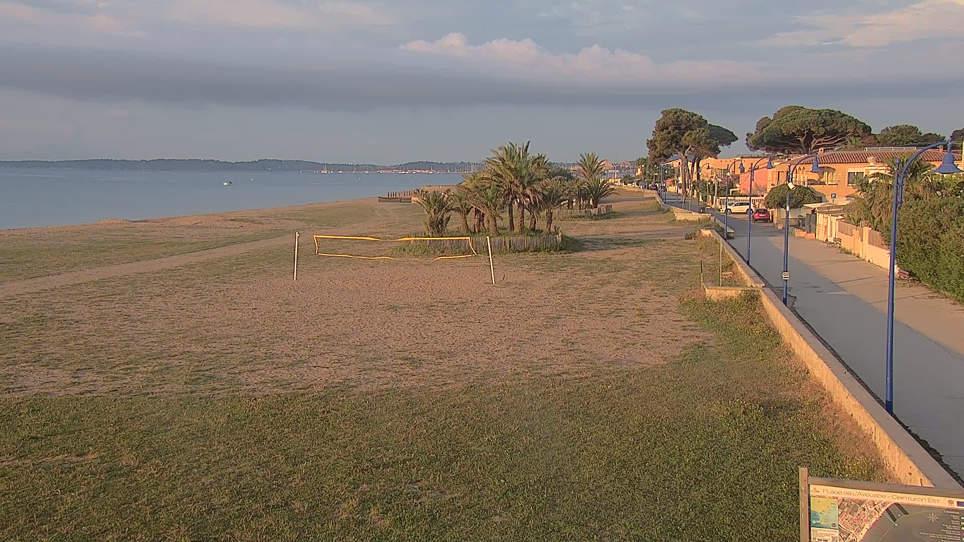A sandy beach with a volleyball net, palm trees, and a paved walkway next to buildings under a partly cloudy sky.