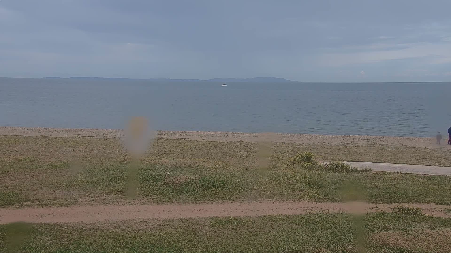 A calm, overcast day reveals a view of a sandy beach, grassy area, and distant land across a body of water.