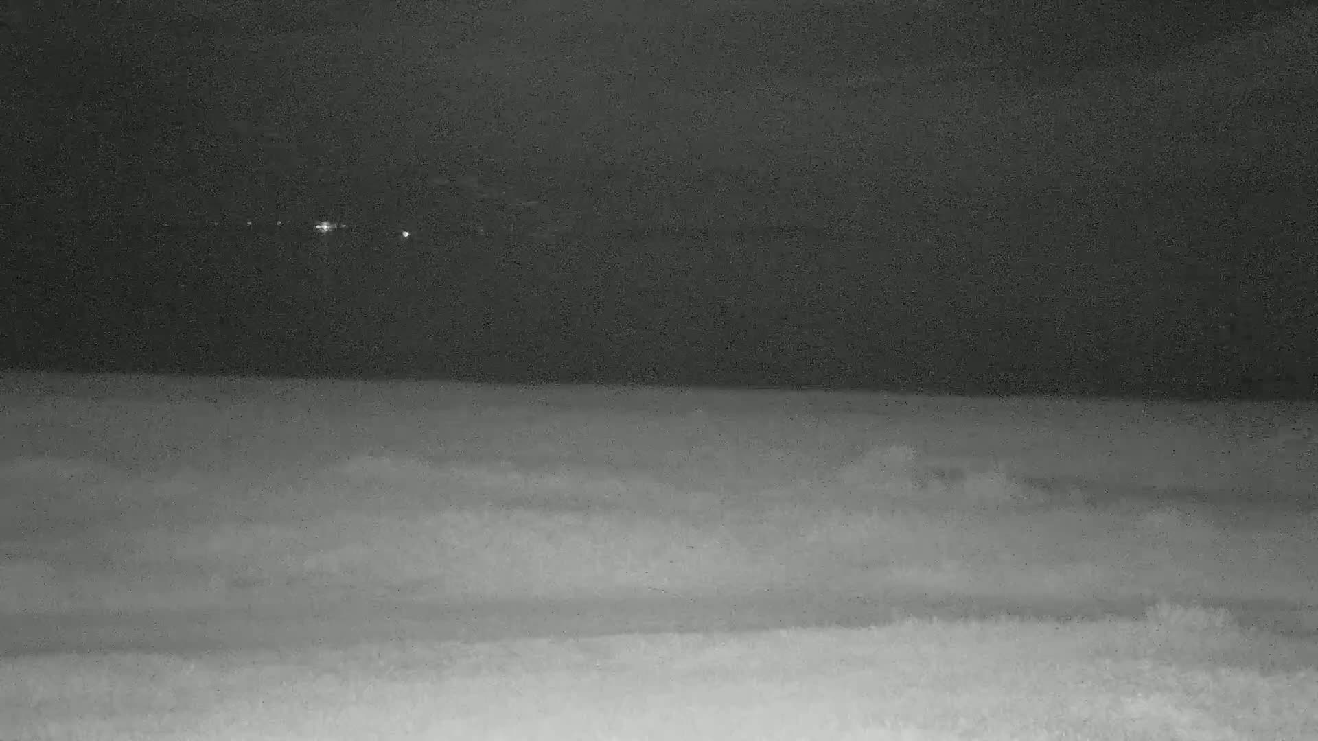 A dark, nighttime scene shows a body of water with distant lights and a flat expanse of land in the foreground.