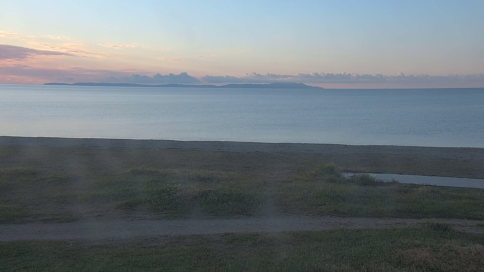 A calm ocean under a pale sunset sky stretches to a distant, low-lying landmass, beyond a sandy beach and grassy area.