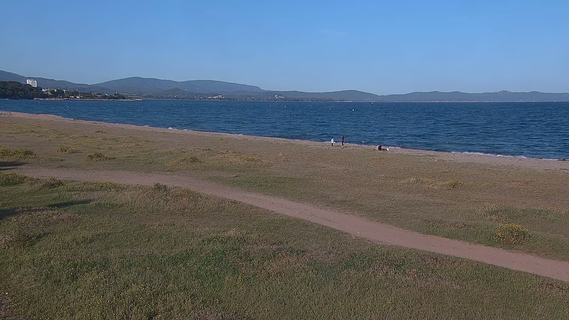 A sandy beach stretches along a calm blue sea under a clear sky, backed by grassy dunes and distant mountains.