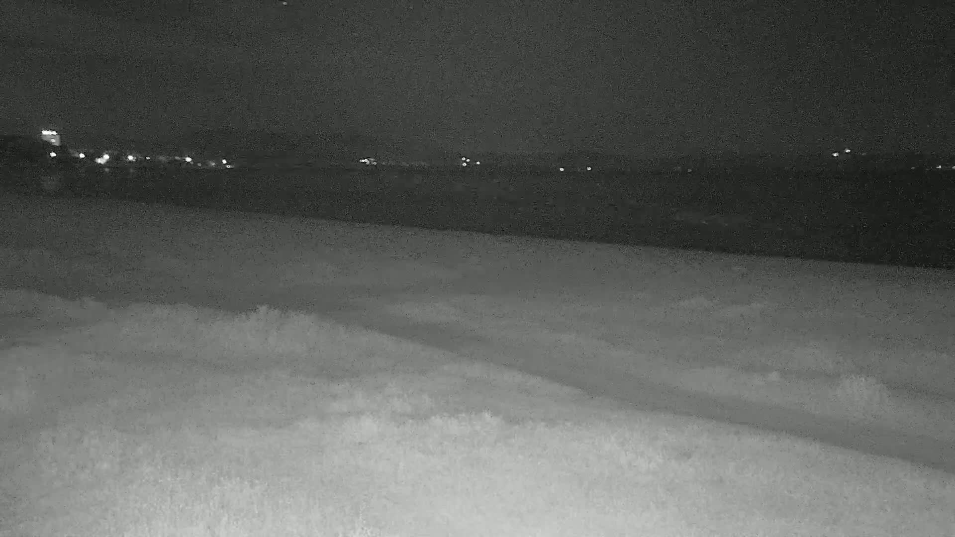 A dark, nighttime scene shows a flat expanse of land with faint lights in the distance across a body of water.