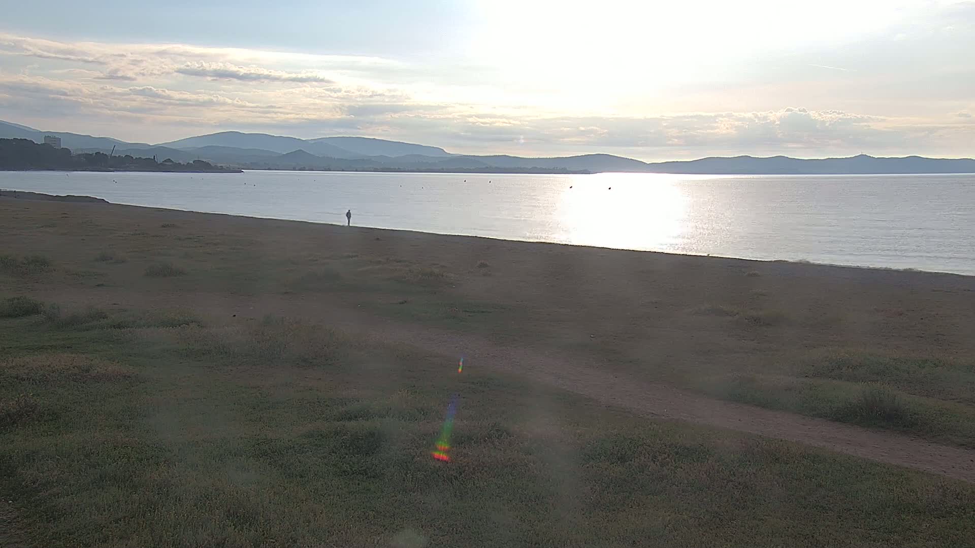 A calm, sunlit body of water stretches to distant mountains under a partly cloudy sky, a sandy beach slopes gently down to the water's edge, with a lone person standing on it.