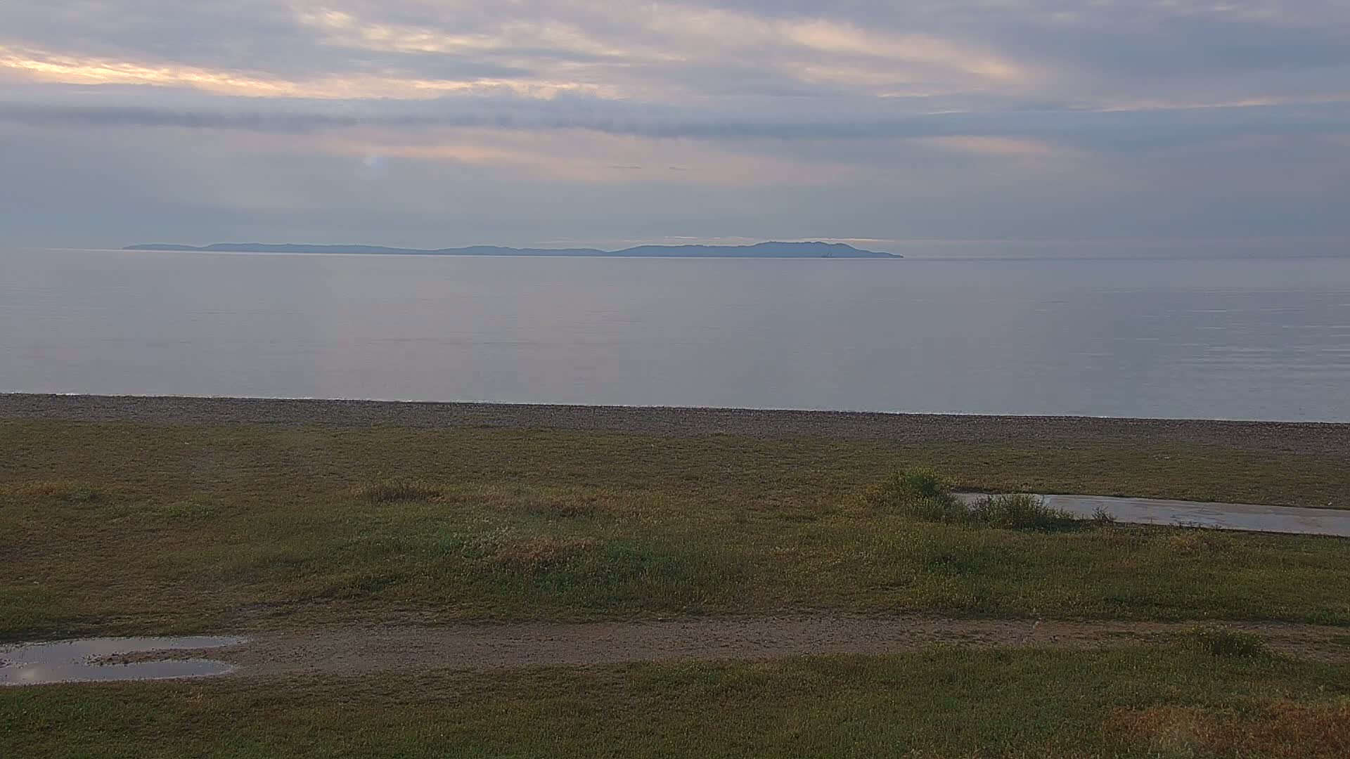 A calm, overcast day shows a long, low island in the distance across a wide body of water, with a grassy area and pebble beach in the foreground.