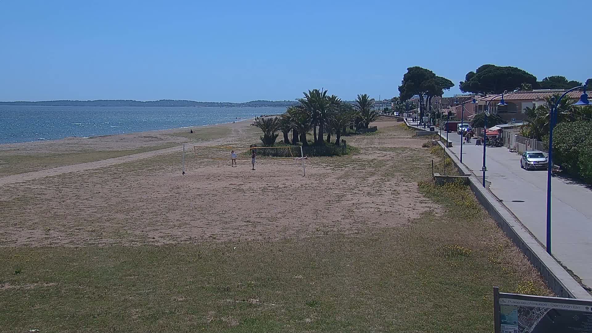 A sandy beach with volleyball nets, palm trees, and a paved walkway beside buildings stretches to a calm, blue sea under a clear sky.