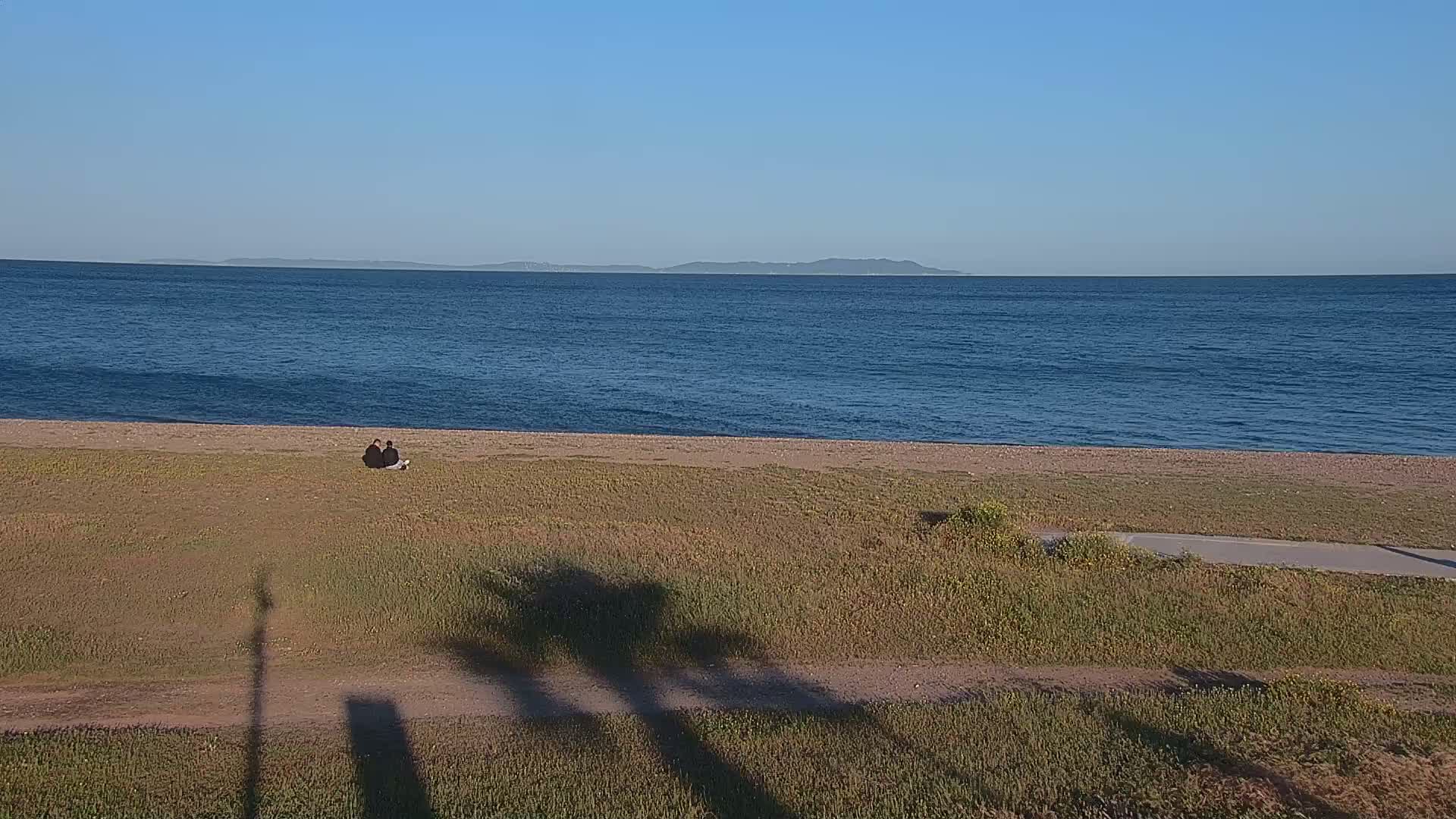 Two people sit on grassy ground near a calm ocean, with a distant island visible under a clear blue sky.
