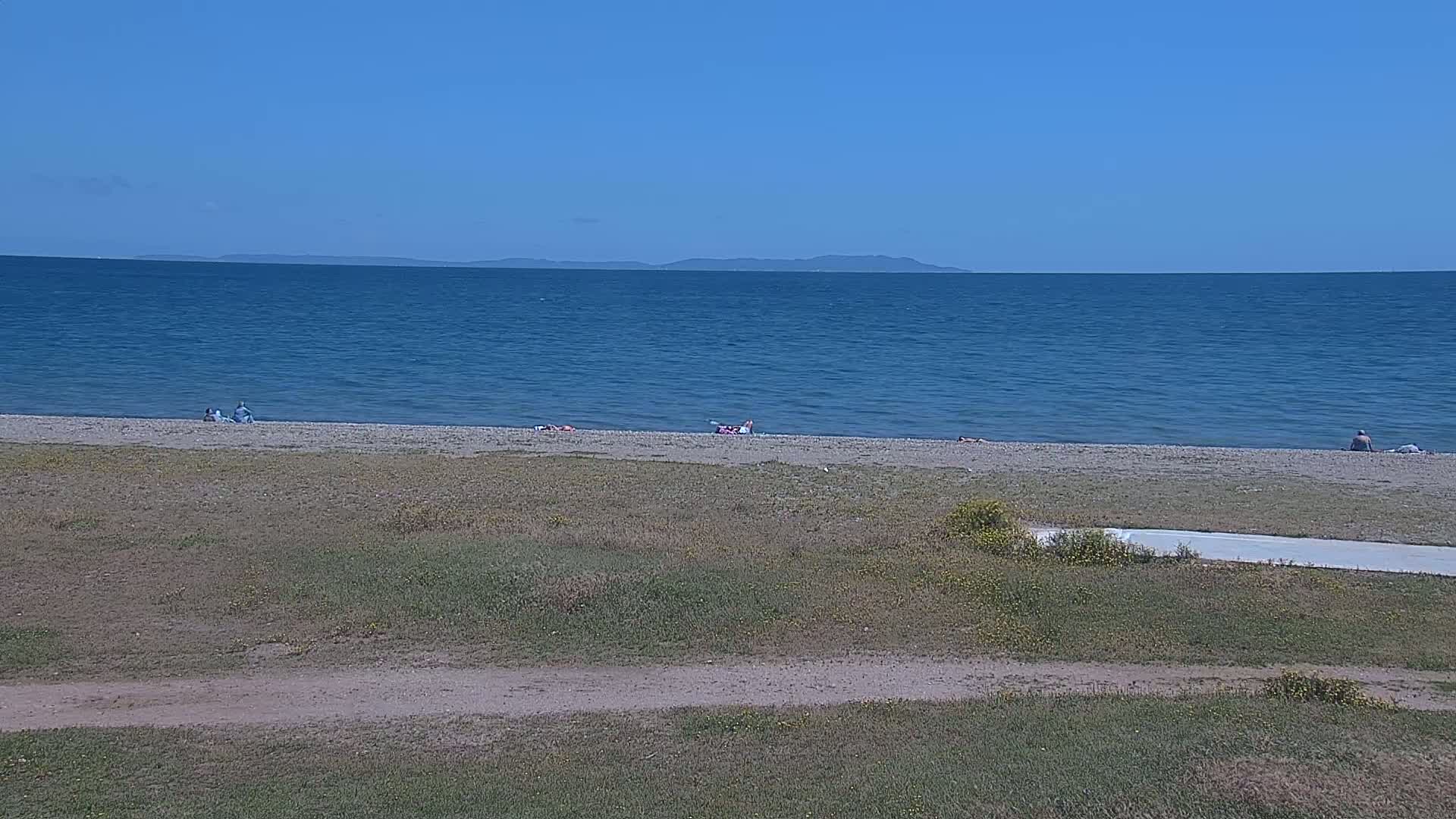 A calm, blue ocean stretches to a distant mountain range under a clear blue sky, with a few people relaxing on a sandy beach in the foreground.