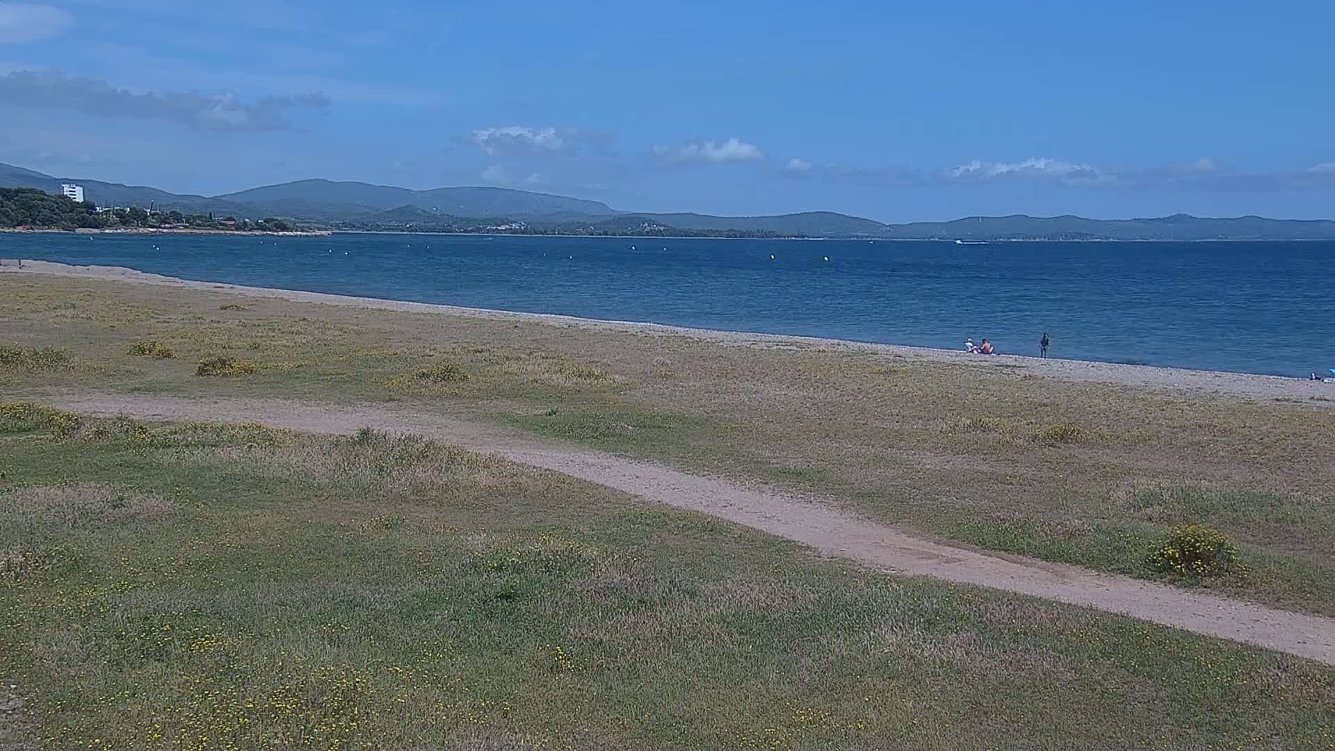 A sandy beach stretches along a calm blue sea under a mostly sunny sky, with sparse vegetation bordering the beach and a dirt path leading toward it.