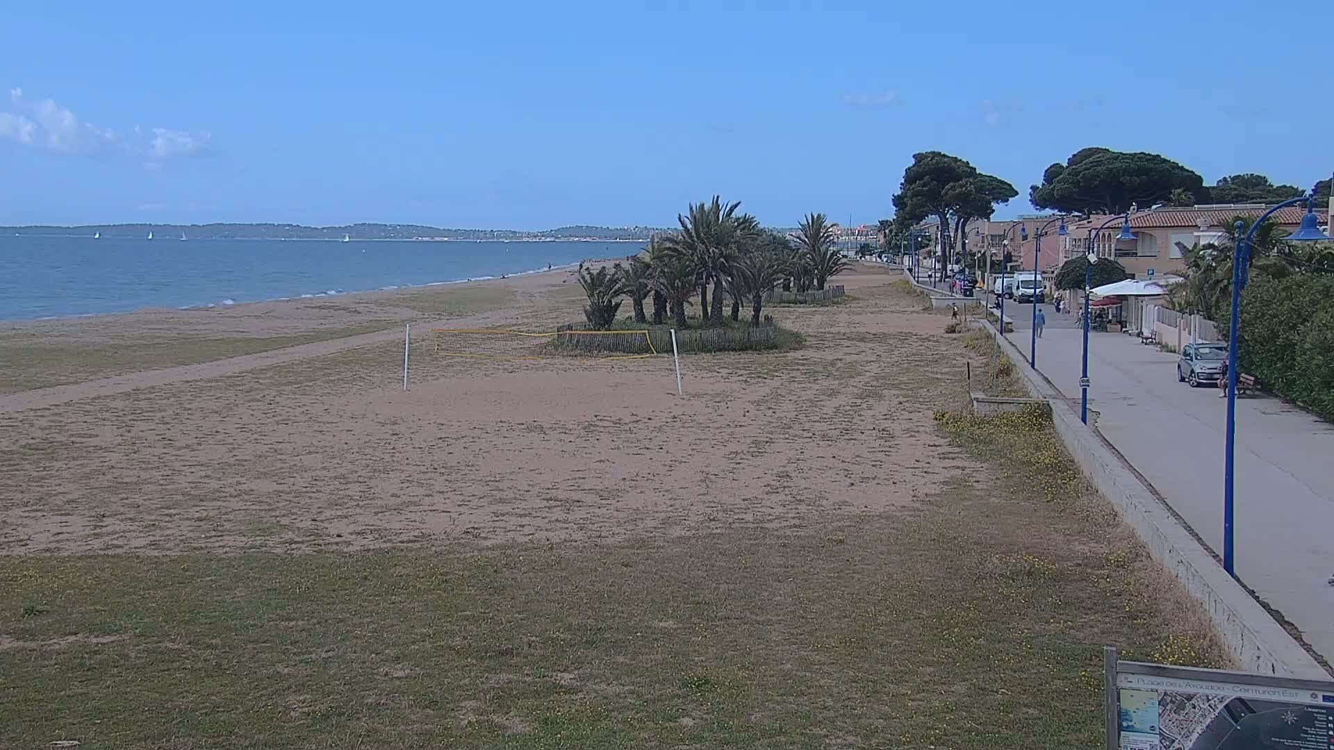 A sandy beach with a volleyball net, palm trees, and a paved walkway bordering buildings stretches along a calm, blue ocean under a clear sky.